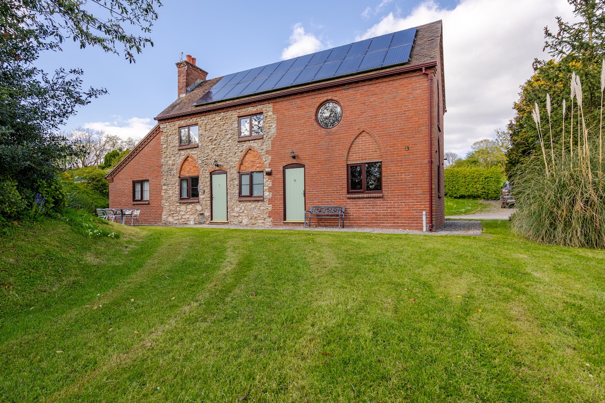 The exterior of a two-story brick and stone cottage is shown, featuring solar panels on the roof. Original architectural details, including arched window and door frames, are visible. A well-maintained lawn surrounds the property, complemented by surrounding greenery.