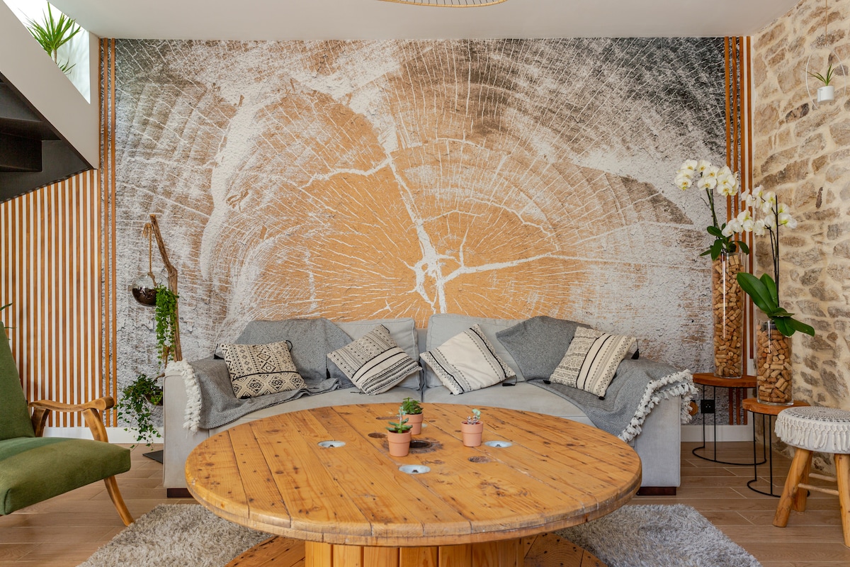 A comfortable living area is highlighted by a large round wooden table, surrounded by a cozy sofa with textured blankets and decorative pillows. A striking wall mural depicting a tree's cross-section adds an organic touch, while potted plants bring a sense of tranquility.