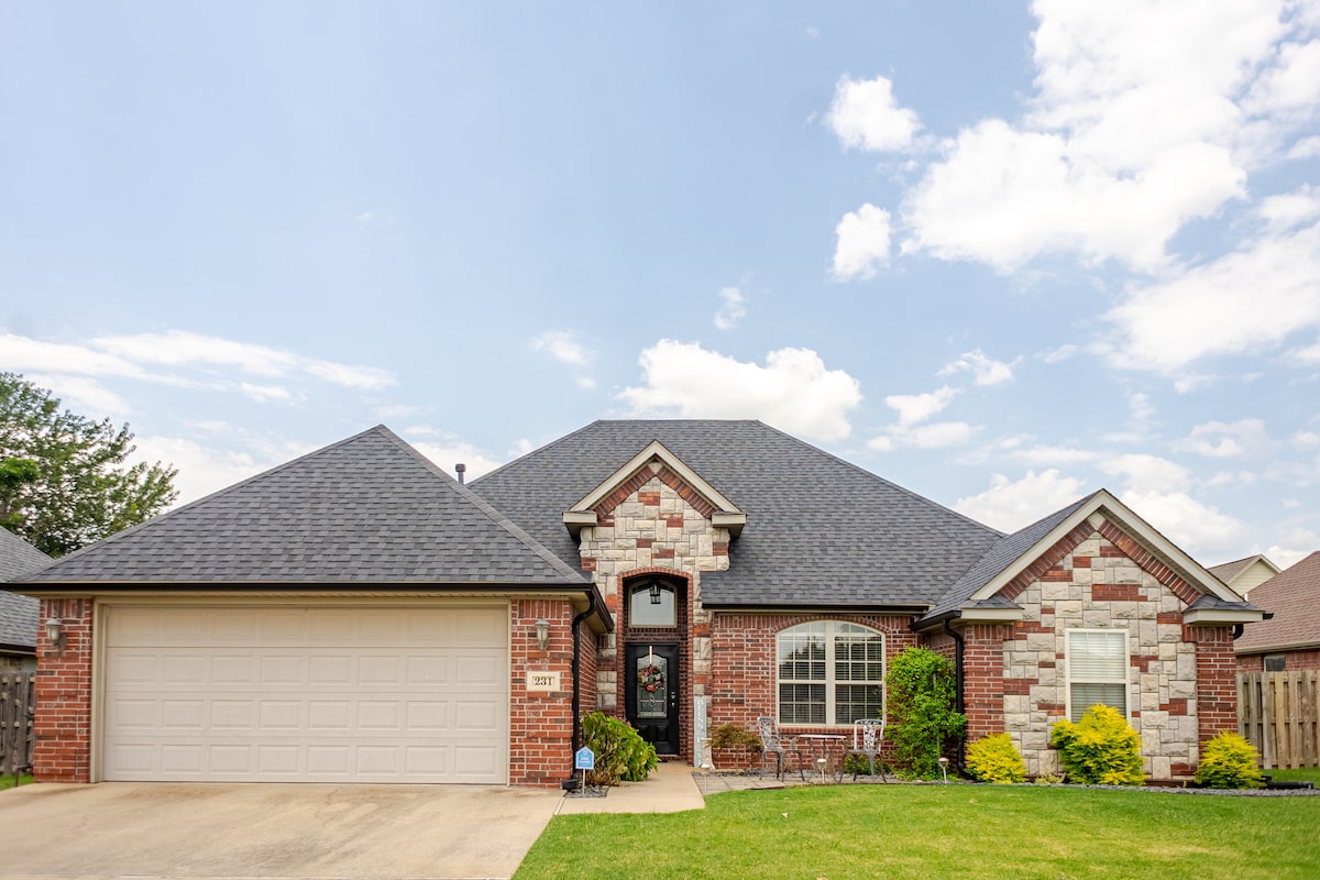 A single-story home is presented with a combination of brick and stone exterior. A spacious driveway leads to a two-car garage. Lush green landscaping enhances the entrance, while large windows welcome natural light into the inviting interior.