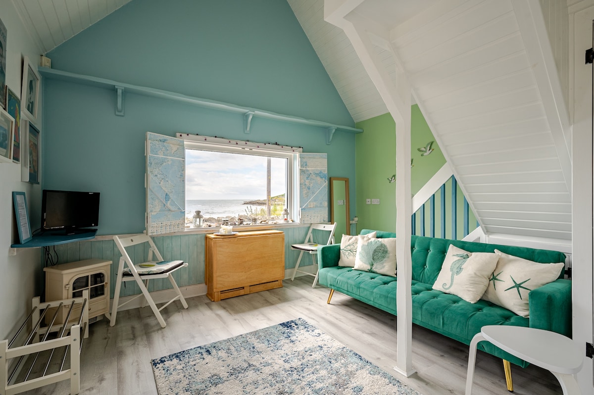 A modern coastal living area features a bright color scheme with aqua and green walls. A view of the beach is framed by large windows, while a comfortable turquoise sofa invites relaxation. A small dining area is visible, along with a television and decorative art.