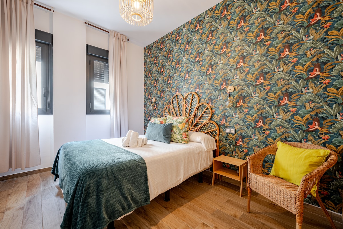 The bedroom features a spacious bed adorned with a green throw and coordinating cushions. A vibrant tropical-themed wallpaper adds color to the room. Natural light filters through two windows with light curtains, while a rattan chair and a small wooden nightstand complete the inviting space.