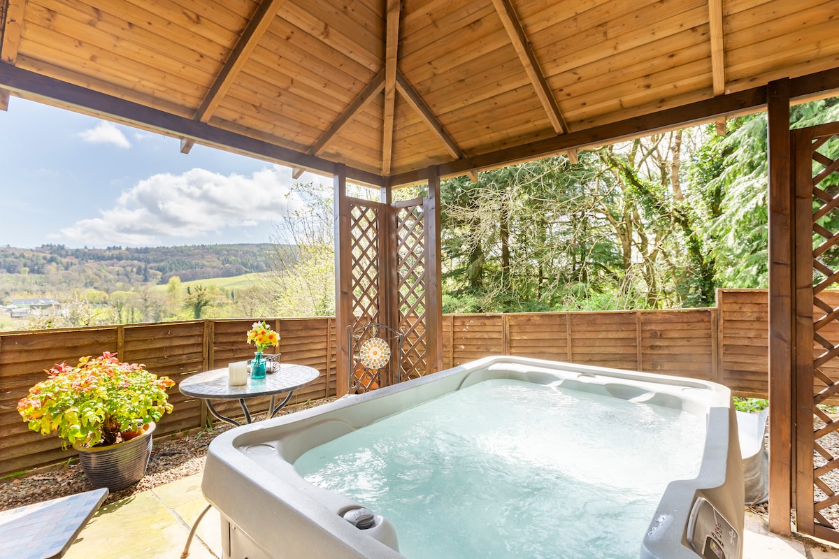 A covered gazebo encloses a spacious hot tub, with vapor rising gently from the surface. Potted plants with vibrant foliage sit nearby, and a round table with two chairs provides a relaxing spot to enjoy the surrounding woodland views.