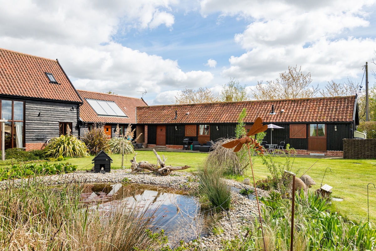 The exterior of the barn features a well-maintained garden with a small pond surrounded by lush greenery. The building showcases a combination of wood cladding and a terracotta tiled roof. Sky-blue clouds drift above, creating a serene countryside atmosphere.