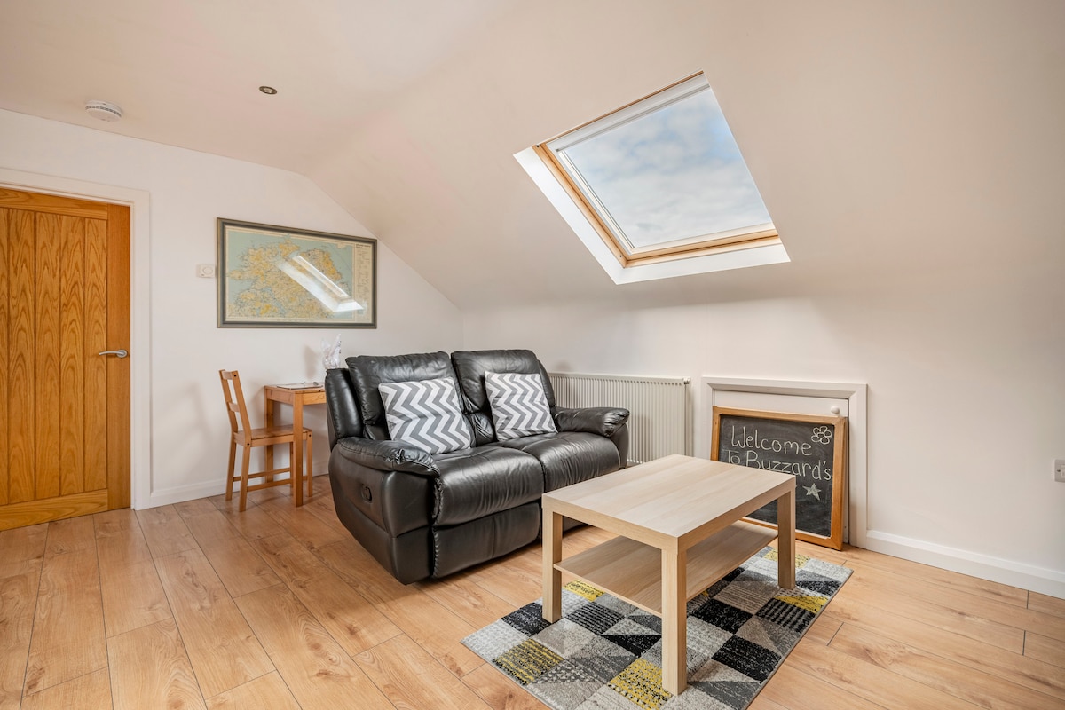 A bright living space features a recliner sofa in front of a small wooden coffee table. Natural light streams through a skylight, illuminating the light wooden floor. A patterned rug adds a touch of color, while a framed welcome message is displayed on the wall.