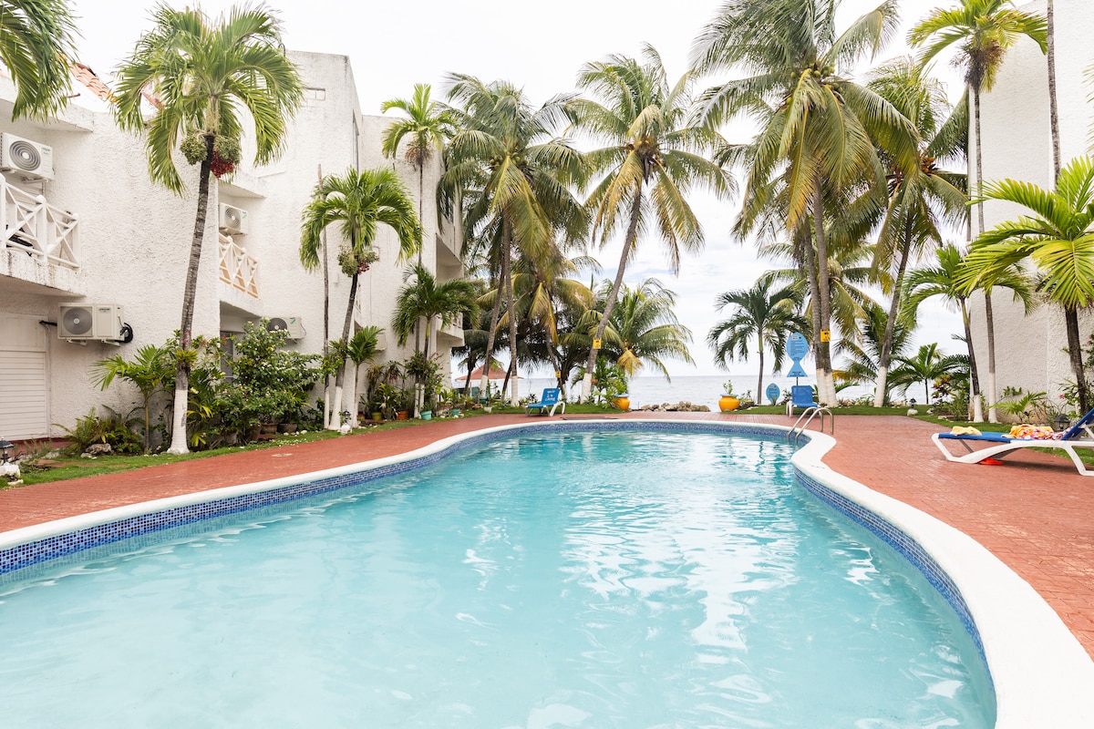 A tranquil swimming pool is surrounded by lush palm trees and manicured gardens. Lounge chairs are positioned nearby, inviting relaxation. The vibrant blue water reflects the cloudy sky, creating a serene atmosphere, with the private beach visible in the background.
