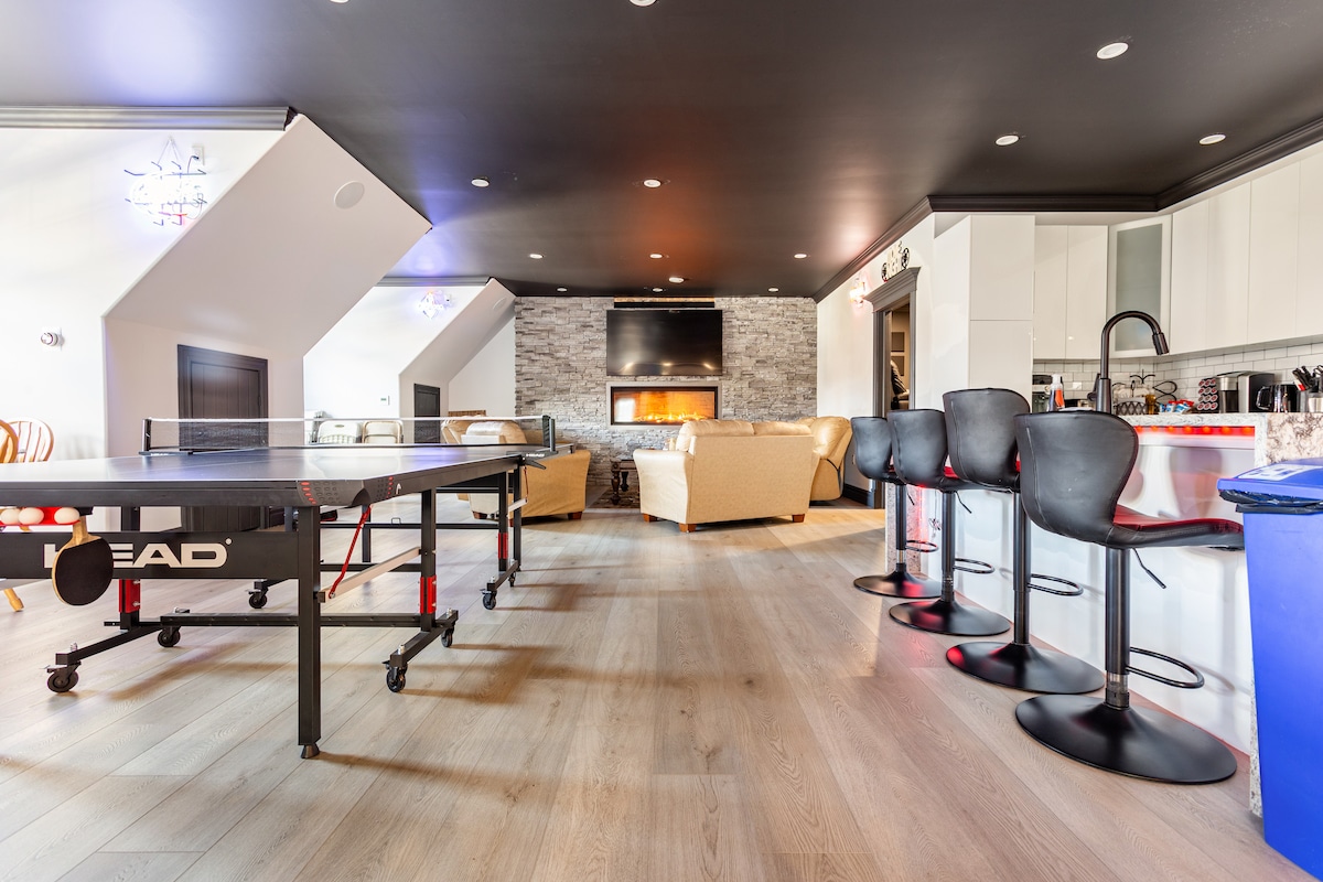 A spacious and modern indoor area features a ping pong table and a cozy seating arrangement. Bar stools line a sleek countertop, while a stone fireplace adds warmth to the space. Natural light fills the area through large windows positioned above.