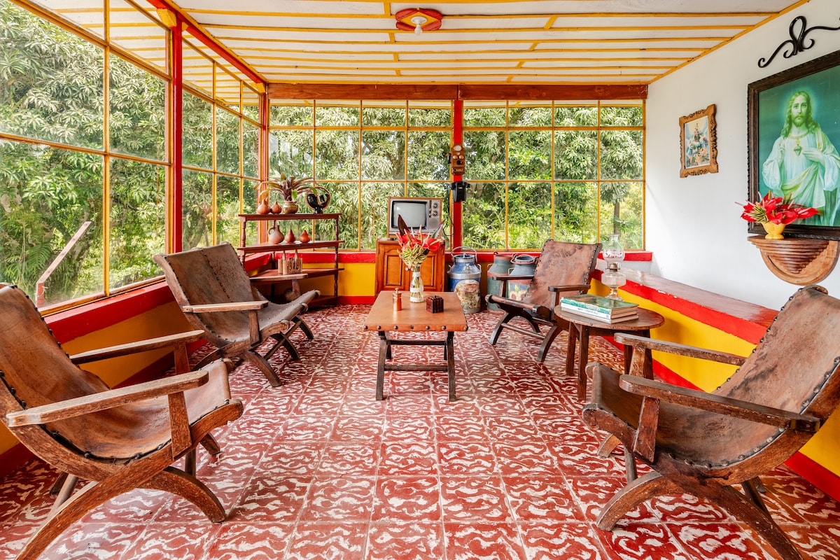 A spacious sitting area is featured, showcasing wooden rocking chairs and a central table. Large windows allow natural light to fill the room, providing views of the surrounding greenery. The floor displays a patterned tile design, complementing the room's vibrant color scheme.