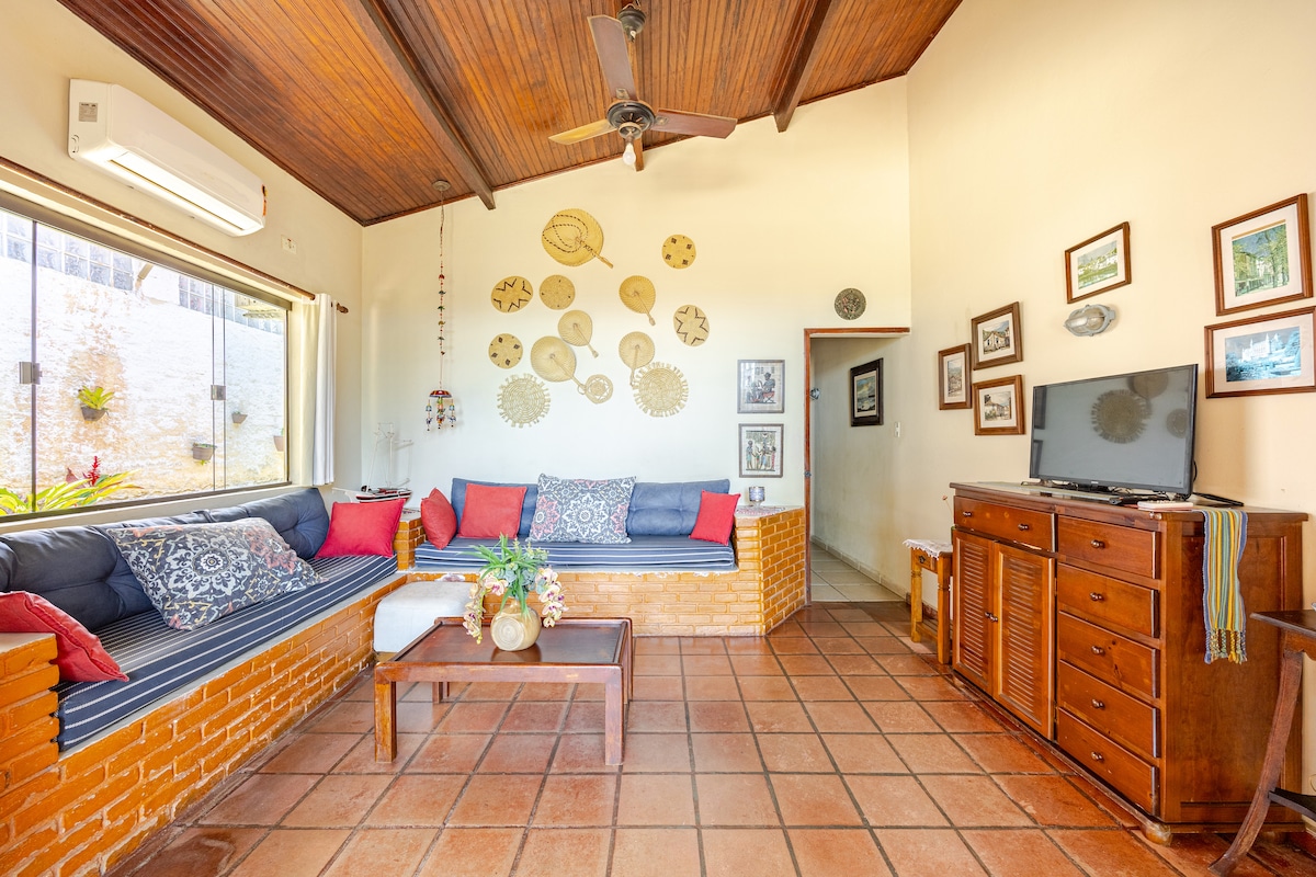 A well-lit living area features a comfortable seating arrangement with decorative pillows on a built-in sofa. A wooden coffee table sits in front, complemented by a sideboard hosting a television. Natural light enters through a large window, enhancing the warm tones of the tiled floor.