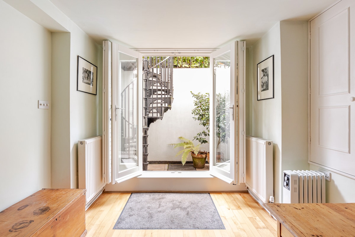 A light-filled living area features large open doors leading to a private patio, enhancing the connection between indoor and outdoor spaces. Two wooden benches are positioned against the walls, with a soft area rug complementing the wooden flooring, and a small potted plant visible in the garden.