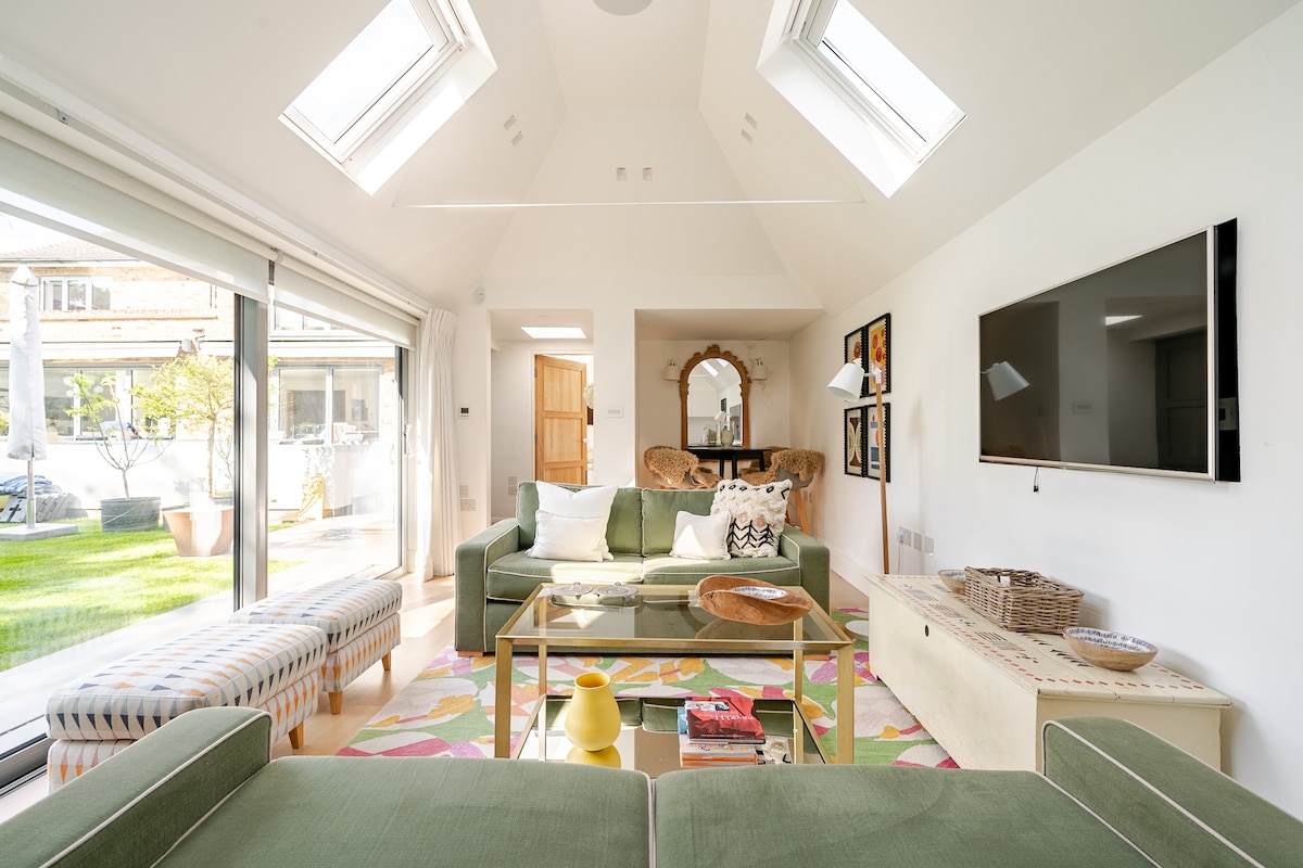 A bright and airy living area is showcased, featuring a spacious green sofa and a glass coffee table. Natural light floods in through skylights and sliding doors, revealing a view of the outdoor space. A colorful rug adds a pop of color to the neutral decor.