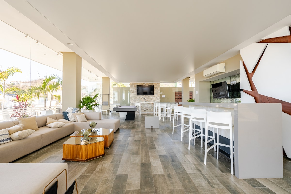 A spacious communal area is illuminated by natural light from large glass windows. Modern furnishings include a plush sectional sofa, stylish wooden coffee table, and high-top white bar stools at a sleek counter. Tropical plants add a touch of greenery to the inviting decor.