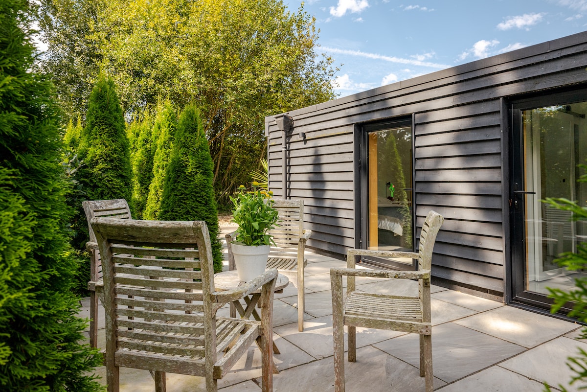 A serene outdoor terrace features wooden chairs arranged around a small potted plant. Lush green hedges frame the space, providing privacy and a natural backdrop. The building's sleek exterior complements the tranquil setting, with glass doors leading into the interior.