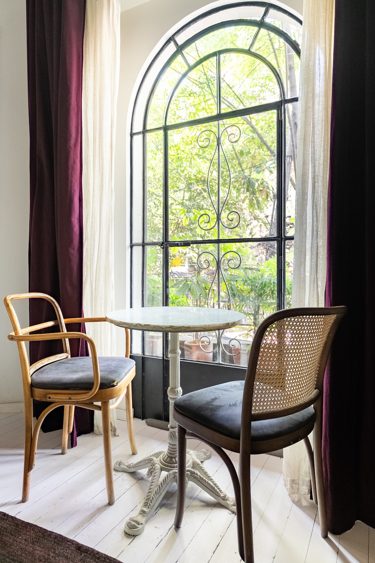 A small round table is positioned between two wooden chairs, one with a woven back. Large windows with elegant metal framing allow natural light to flow in, offering views of the green foliage outside, while sheer curtains softly frame the scene.