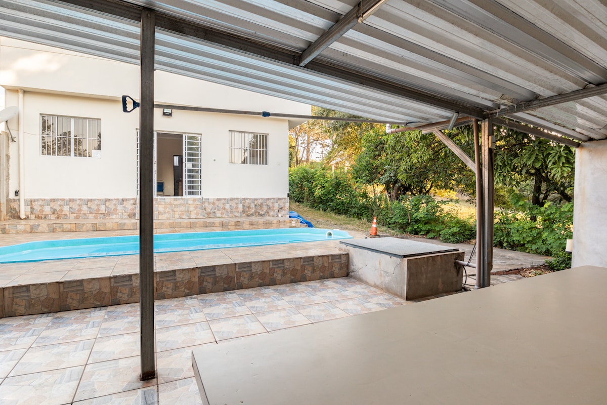 An outdoor area is presented with a spacious terrace featuring a stone border and a large table. A solar-heated pool is visible in the background, surrounded by greenery, with a sliding door leading into the house.