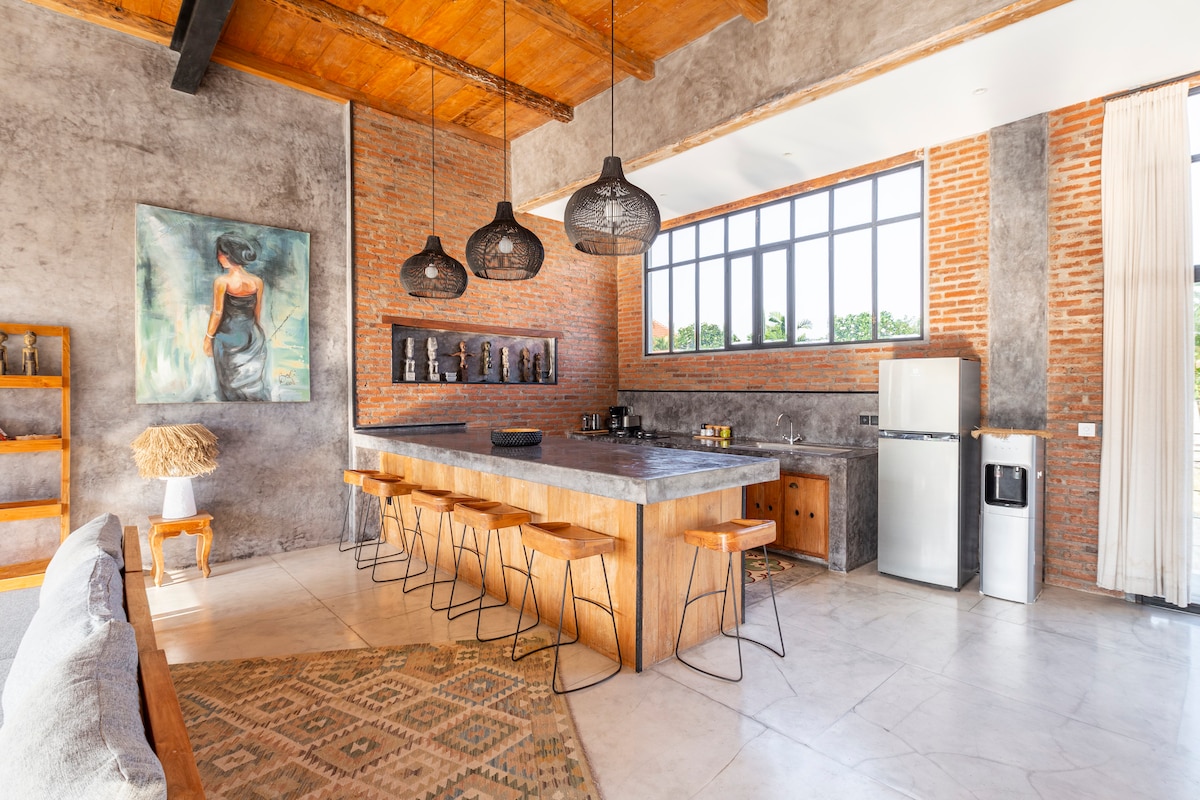 The open-concept kitchen features a large concrete island with six wooden stools, designed for casual seating. Exposed brick walls complement the warm wooden beams overhead. Natural light floods the space through large windows, illuminating the modern appliances and stylish decor.
