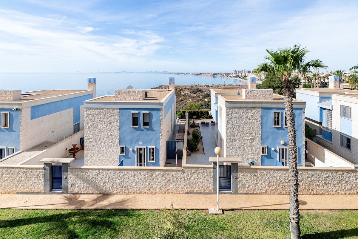 A view of a coastal apartment complex featuring blue and beige buildings. Lush greenery is present in the foreground, complemented by palm trees. The sea is visible in the background, extending towards the horizon under a partly cloudy sky.