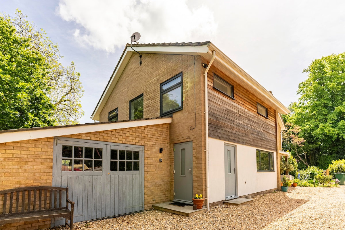 A modern guest annexe features two distinct exterior textures, with a combination of brick and wood accents. Natural light reflects off large windows, and a gravel driveway leads to the front doors. Lush greenery surrounds the property, adding to the peaceful environment.
