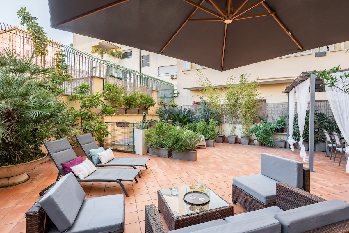 The spacious terrace features comfortable seating with a low table, surrounded by lush greenery in planters. A large umbrella provides shade, and sun loungers are positioned for relaxation. The warm terracotta tiles enhance a welcoming outdoor space for gatherings or quiet moments.