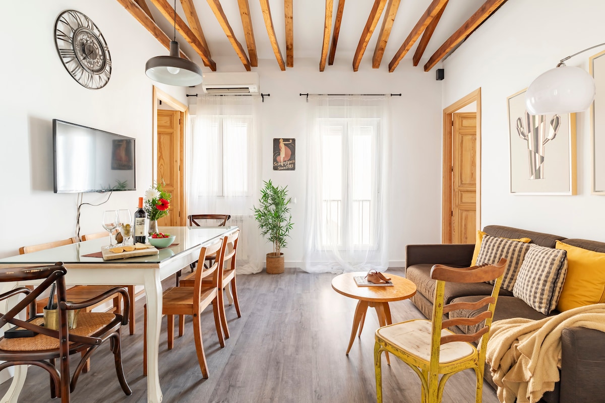 A spacious living area features a modern sofa with yellow cushions, a wooden coffee table, and a dining table surrounded by chairs. Natural light streams through two windows, and wooden beams enhance the ceiling's character, contributing to a bright and inviting atmosphere.
