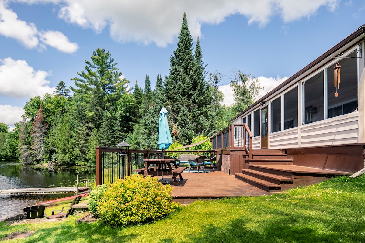 A spacious outdoor deck is presented, featuring a picnic table and umbrella, surrounded by lush greenery. A pathway leads to a private dock on the water, framed by tall trees and a clear blue sky.