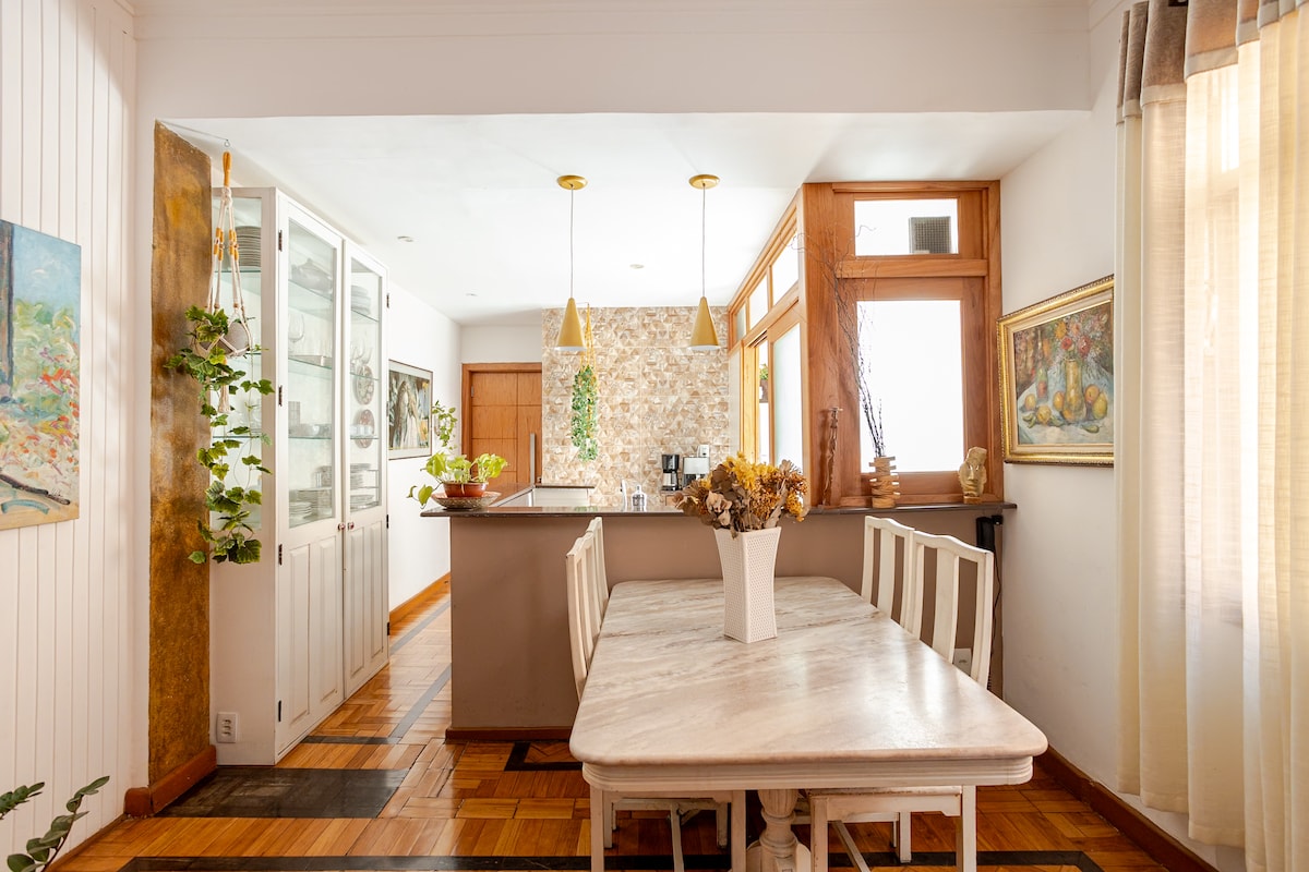 A dining area is presented with a marble-top table surrounded by light-colored chairs. Natural light filters through large windows, illuminating the space. A partial view of the kitchen showcases modern fixtures and decorative plants, adding a refreshing touch to the warm ambiance.