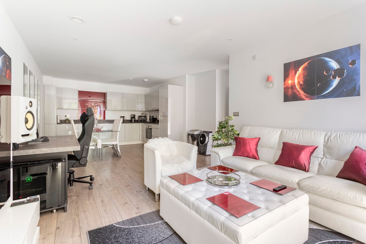 A modern living room features a large white sectional sofa adorned with red accent pillows. A glass coffee table sits in the center, while a high-quality speaker and an office chair are positioned nearby. The open kitchen area is visible in the background, enhancing the spacious layout.