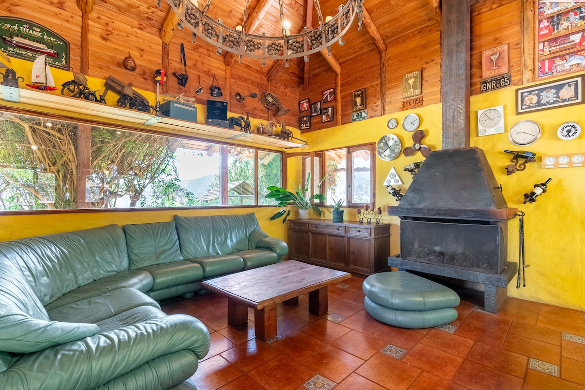 A spacious living area is characterized by a high wooden ceiling and vibrant yellow walls. A large, curved green couch surrounds a wooden coffee table. A stone fireplace and assorted wall decorations add to the room's rustic charm. Ample natural light filters through the windows.