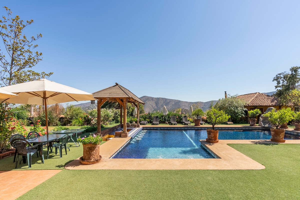 A spacious outdoor area is centered around a large swimming pool, framed by greenery and potted plants. A gazebo offers shaded seating, and a dining table with chairs invites gatherings. The backdrop features rolling hills under a clear blue sky.