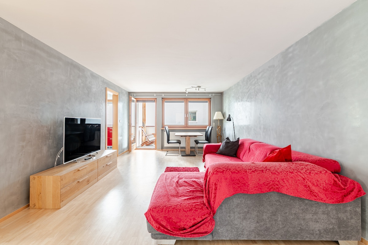 A spacious living room features a large red sofa and a light wooden TV stand with a television. Nearby, a dining area is visible with a sleek table and chairs. Natural light fills the room through large windows, creating a bright and open atmosphere.