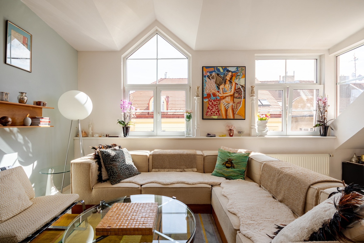 A spacious living area is highlighted by a large corner sofa with cozy throw pillows. Sunlight enters through angled windows, illuminating a glass coffee table and a textured rug. A colorful art piece adorns the wall, complemented by decorative items on a shelf.