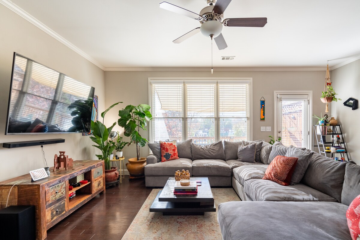 A spacious living area features a large sectional sofa adorned with colorful cushions. Natural light pours in through multiple windows, complemented by a vibrant potted plant. A wall-mounted television and a decorative wooden console add functionality and warmth to the space.