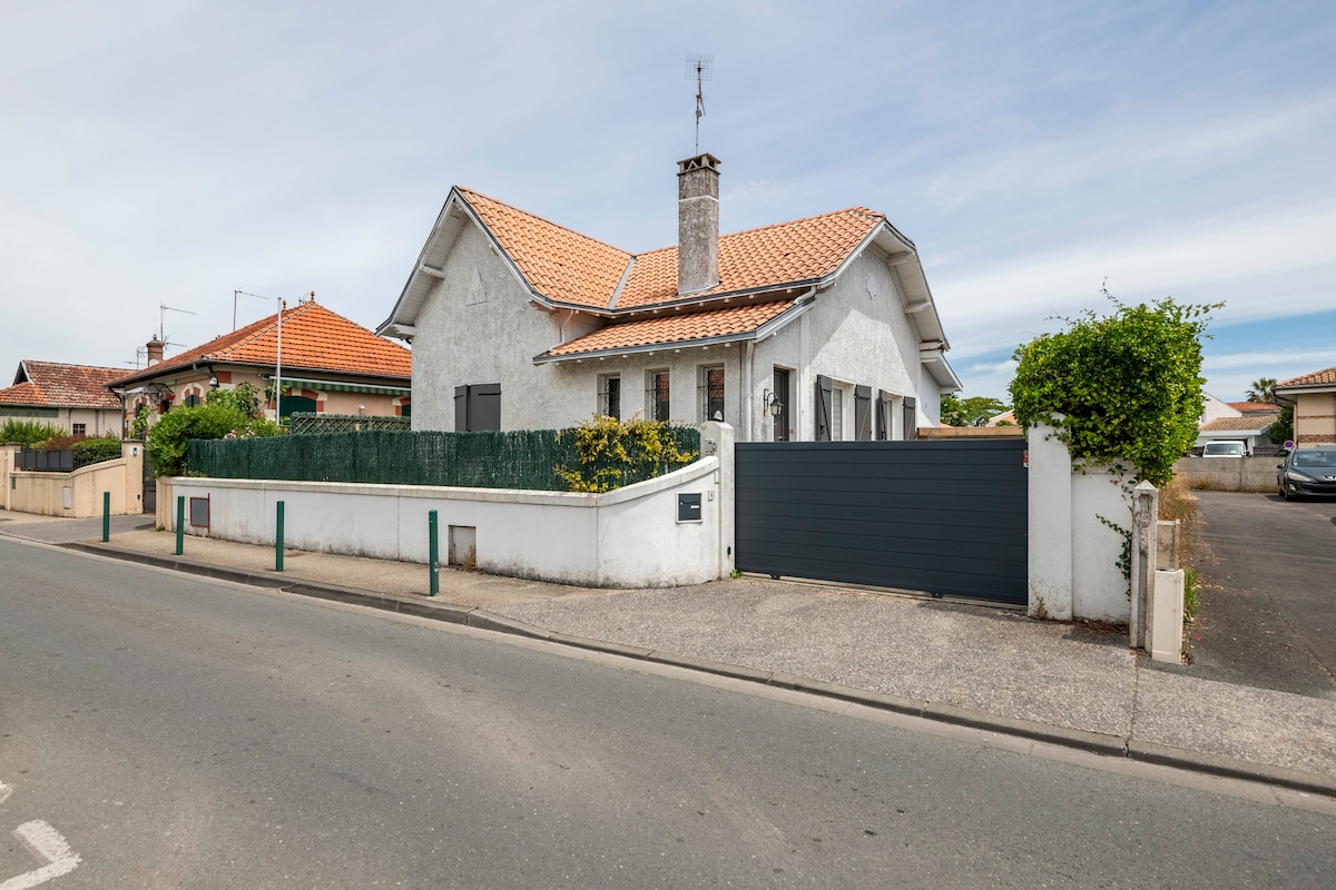 The exterior of a traditional house is depicted, featuring a tiled roof and a stone facade. A gray gate leads to the entrance, bordered by greenery and a low fence. The paved pathway invites guests towards the front door, emphasizing the property’s welcoming appeal.