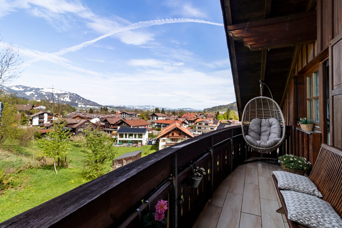 A balcony is displayed, featuring a hanging chair and a wooden bench. The panoramic view reveals charming village houses and lush greenery beneath a clear blue sky striped by contrails, with distant mountains providing a scenic backdrop.