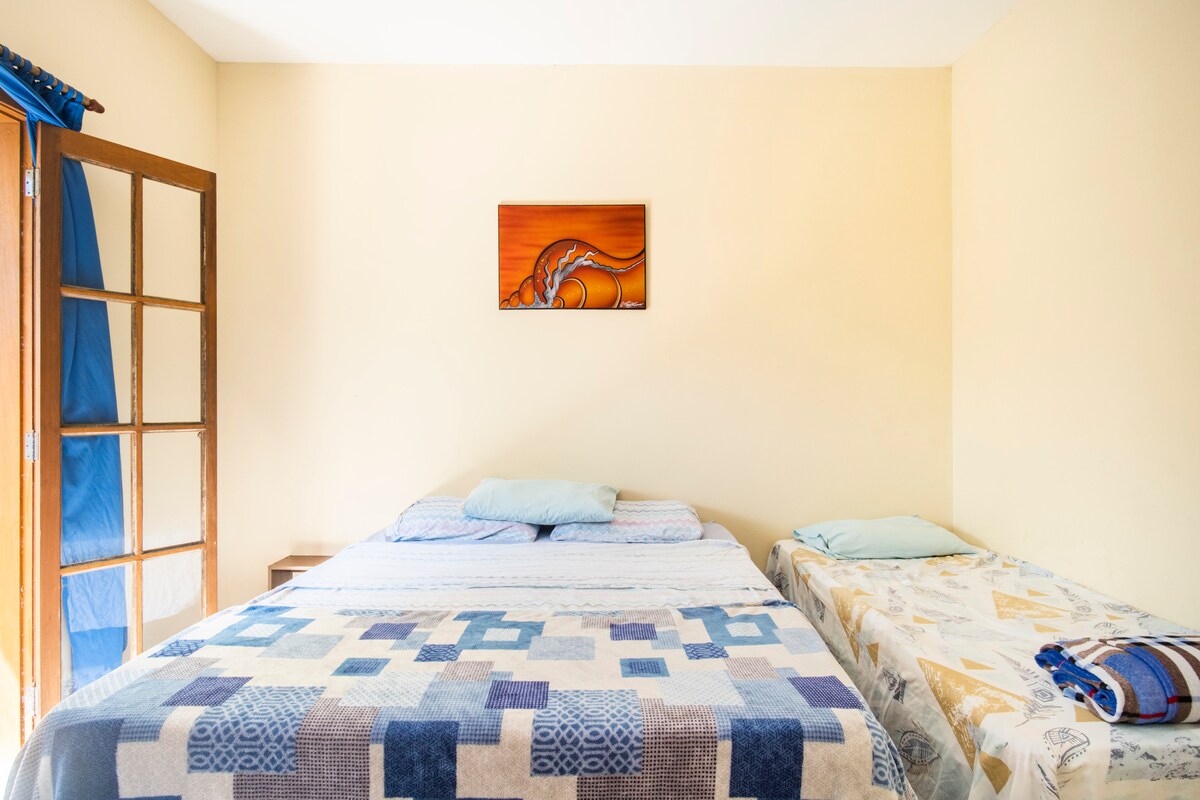 A cozy sleeping area is presented with a spacious queen-size bed and a single bed, both topped with soft bedding. A piece of wall art adds a touch of color, while natural light enters through nearby glass doors, illuminating the room's warm tones.