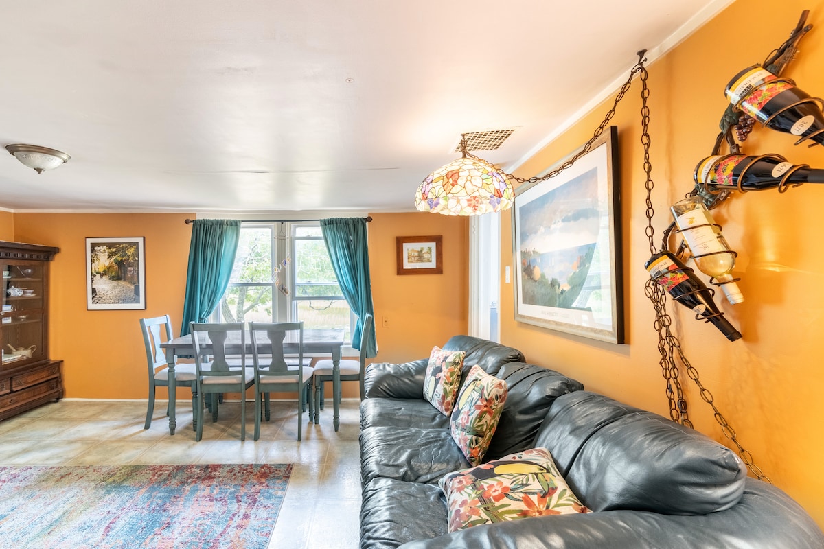 A comfortable living area features a dark leather sofa with decorative pillows, paired with a dining table and chairs. Warm orange walls are complemented by large windows dressed in blue curtains, allowing natural light to brighten the space. A unique wine display adorns the wall.