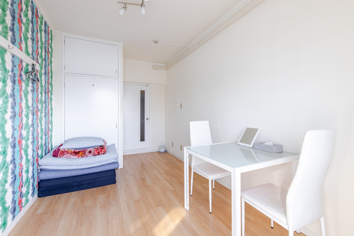 A bright and airy room features a wall decorated with green and blue patterns. A white table is positioned with two chairs, while futon beds are neatly arranged on the floor. A closet and door are visible, contributing to the room's functionality.