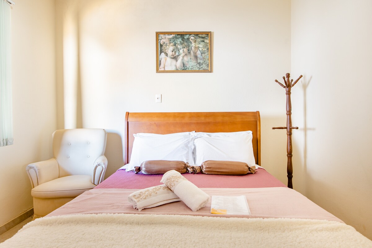 A well-lit sleeping area is equipped with a spacious bed adorned with neutral-colored linens. A cozy armchair is positioned nearby, complemented by a decorative wall art piece. A coat rack stands against the wall, adding to the room's functionality.