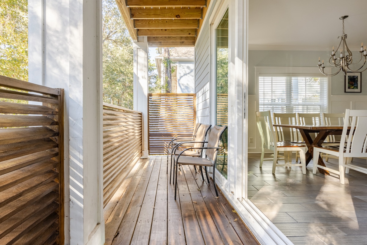 A covered porch features two chairs positioned on wooden decking, providing a space for relaxation. Sunlight filters through nearby trees, casting gentle shadows. The open interior is visible through large glass doors, showcasing a dining area with a wooden table and white chairs.