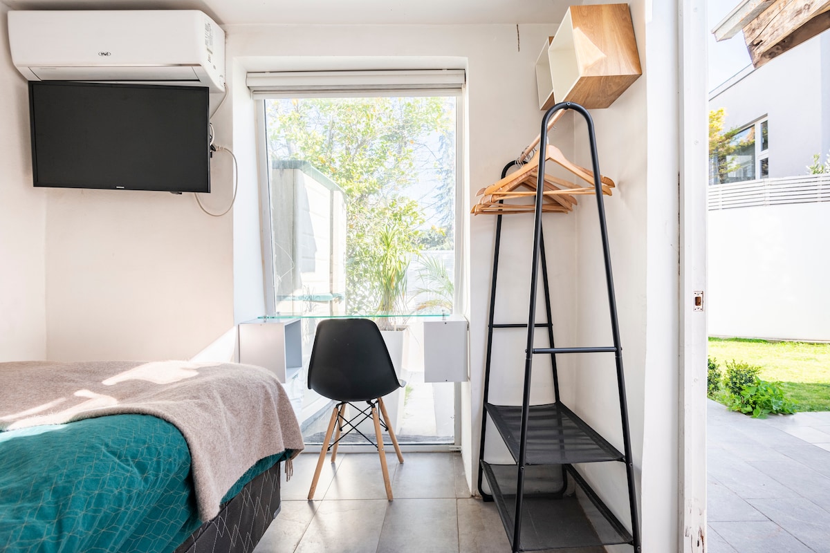 A small, cozy room is presented, featuring a comfortable bed with a soft blanket. Natural light filters through a large window, enhancing a serene atmosphere. A modern chair and a wooden shelf add functionality, while a black storage unit provides additional space for personal items.