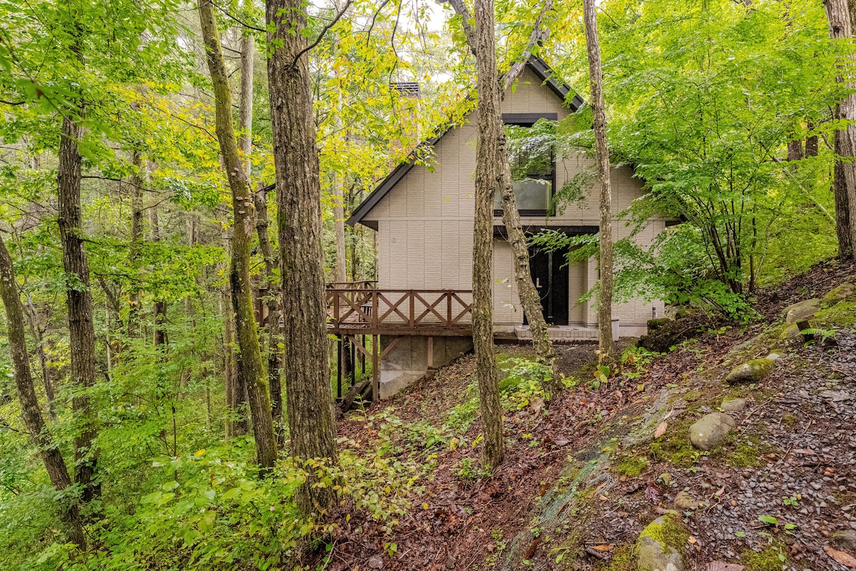A two-story cottage is nestled among lush greenery, framed by tall trees. A wooden deck extends from the home, providing an inviting entrance. The natural surroundings create a serene environment, complementing the structure's modern design.