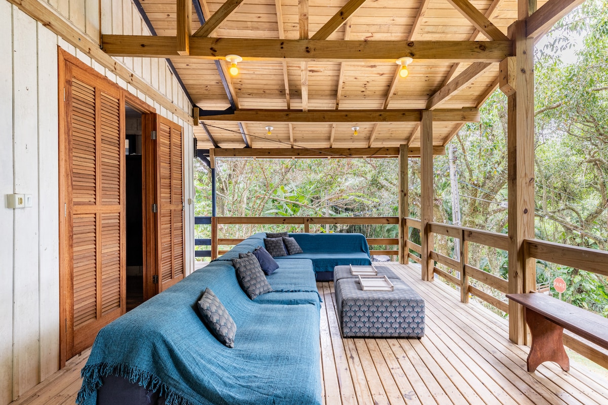 A spacious covered terrace features comfortable seating arrangements with soft, textured fabrics. Wooden floors and structural beams provide a rustic feel, while surrounding greenery adds a natural touch. Large open spaces allow for ample natural light and fresh air.
