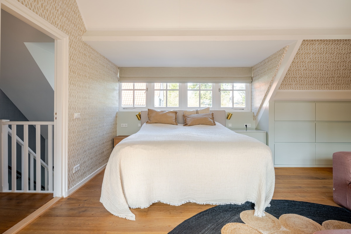 A spacious sleeping area features a large bed covered with a textured white throw. Two bedside lamps provide soft lighting, and natural light filters through multiple small windows along the wall. A stylish rug adds warmth to the wooden flooring.