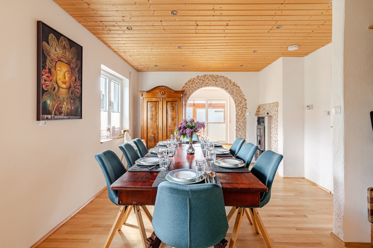 A spacious dining area is featured, with a long wooden table set for dinner. Colorful chairs surround the table, complementing the light wood ceiling and bright walls. Natural light enters through large windows, illuminating an elegant sideboard and a decorative wall art piece.
