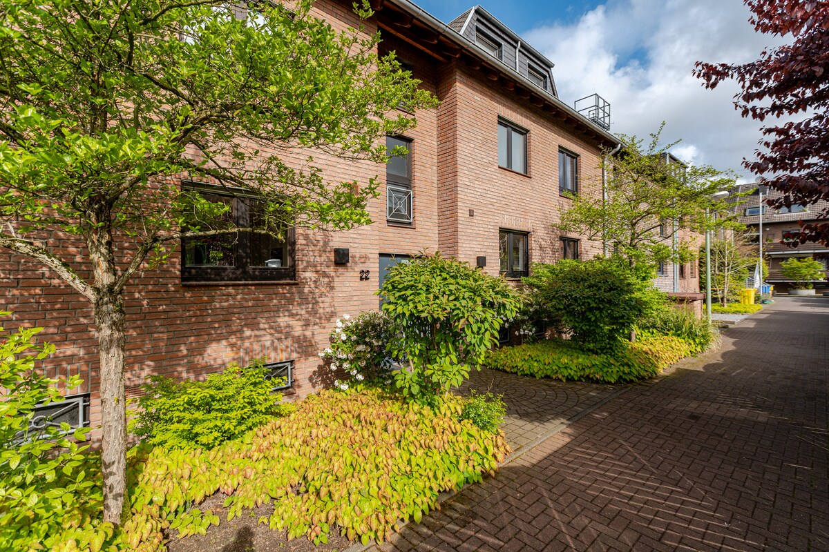 The building's brick facade is complemented by lush green foliage and colorful plants lining the pathway. Large windows reflect natural light, enhancing the inviting exterior. The setting is framed by well-maintained trees, creating a peaceful atmosphere near the entrance.