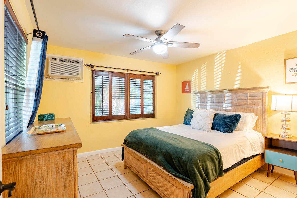 A bright bedroom features a spacious bed adorned with soft linens. Natural light filters through partially opened shutters, highlighting the warm yellow walls. A ceiling fan provides airflow, and an air conditioning unit is installed nearby, ensuring comfort in the room.