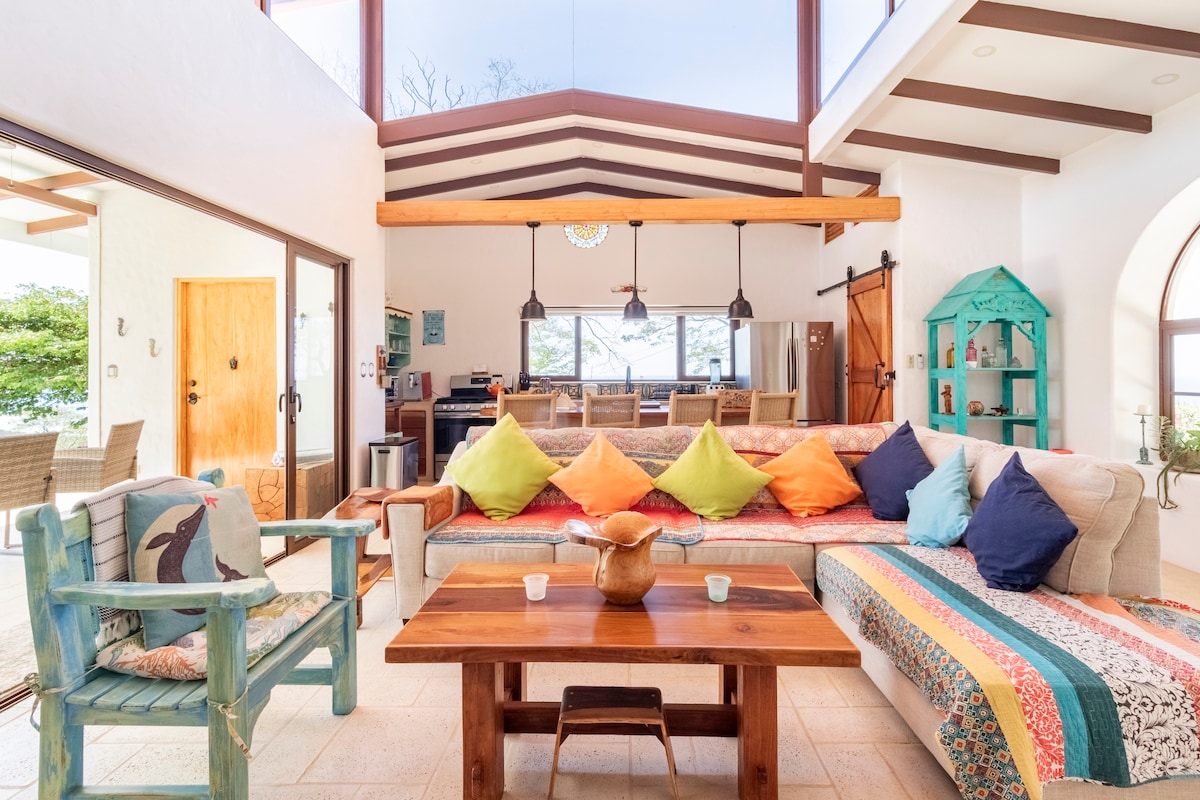 The bright living area features high, vaulted ceilings and large windows allowing natural light. A colorful assortment of cushions decorates a spacious sofa, complemented by a wooden coffee table in front. A small, turquoise cabinet and dining space are visible in the background.