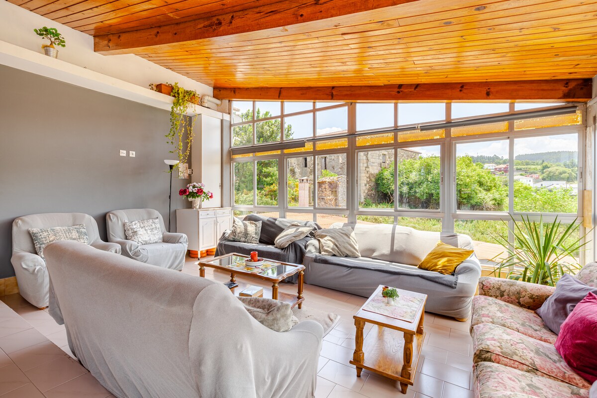 A spacious living area features large windows, allowing natural light to illuminate the room. Comfortable seating is arranged around a low table, with a mix of soft sofas and chairs. Greenery is visible outside through the windows, enhancing the serene atmosphere.