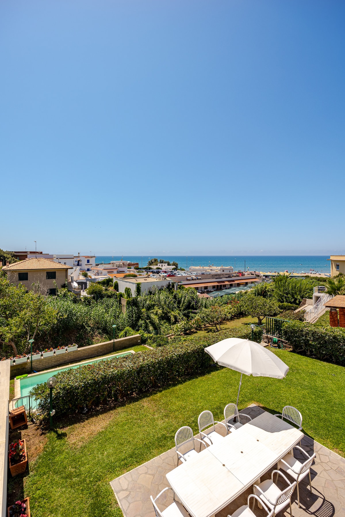 A view from a garden reveals a large outdoor dining table surrounded by white chairs. Green grass and hedges frame the setting, leading to the coastline in the background. A bright blue sky complements the vibrant scene, while an umbrella provides shade over the table.