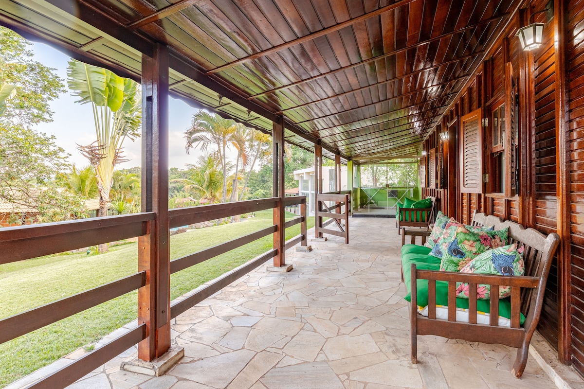A covered porch features wooden beams and stone flooring, offering comfortable seating with lush green cushions. The area is surrounded by greenery, with palm trees visible in the background, creating a peaceful outdoor space for relaxation.