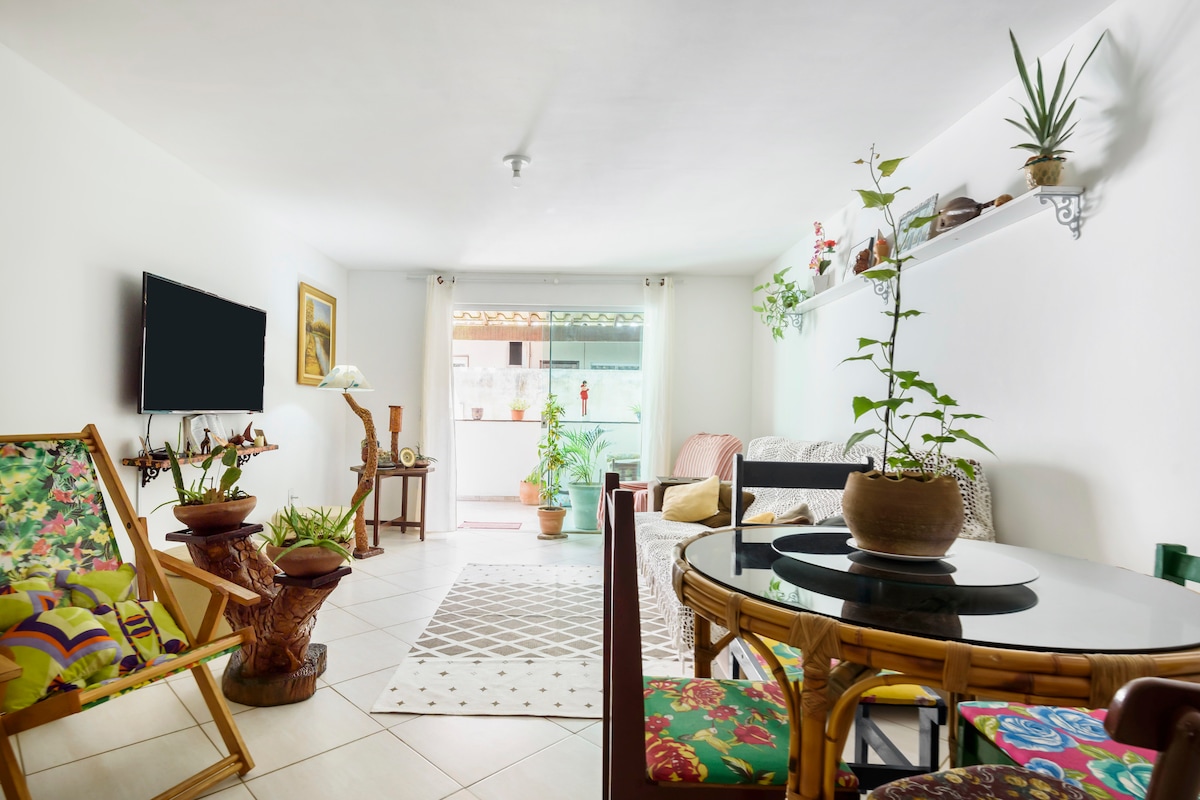 A bright living space features a comfortable seating area with floral-patterned chairs and a cozy couch. A round table sits in the center, adorned with a plant. Light filters in through a window, highlighting decorative shelves and potted greens along the walls.